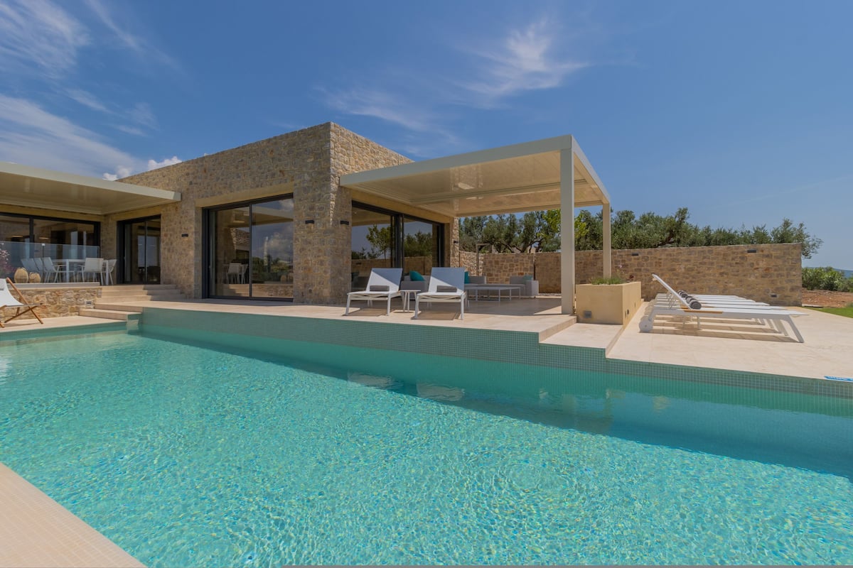 Ein moderner Poolbereich mit Liegestühlen, einer überdachten Lounge und einem Steinhaus mit großen Fenstern.