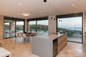A modern kitchen with a large island, dining area, and large windows offering a view of the landscape. - Messinian Nest (Pylos-Nestoras)