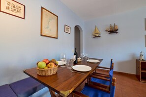 A dining area with a wooden table set for two, a basket of fruit, and framed pictures on the wall. - Sunny Haven at the Village Square (Palafrugell)