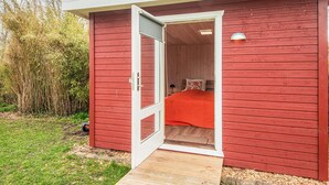 Eine rote Holzhütte mit einem Einzelbett, einer kleinen Veranda und einer Tür zum Schlafzimmer.