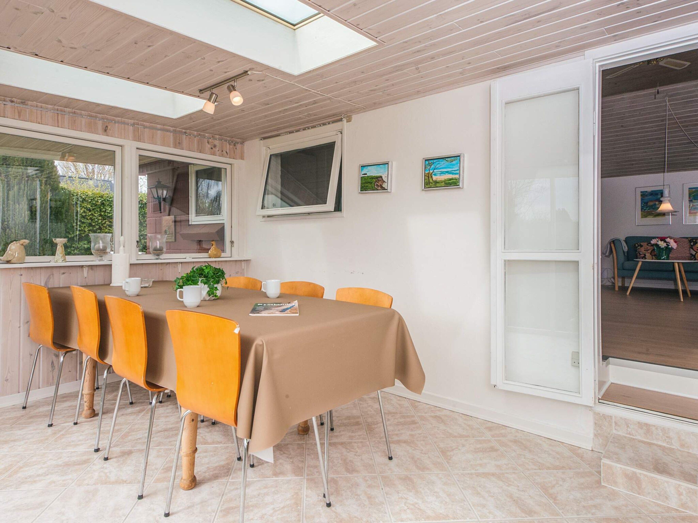 A dining area with a table set for six, surrounded by orange chairs, with a large window and artwork on the wall.