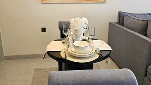 Uma sala de jantar com uma mesa posta para dois, um quadro na parede e um sofá cinza ao fundo.