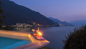A lakeside terrace with a pool, candles, and a view of a mountainous town at dusk. - Villa Meraviglia  (Olgiasca)