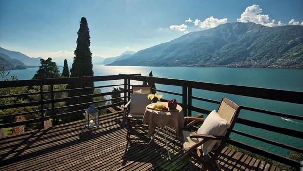 A wooden deck with a table and chairs overlooking a lake with mountains in the background. - Villa Meraviglia  (Olgiasca)