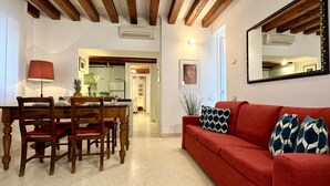 Ein Essbereich mit einem Holztisch und Stühlen, einem roten Sofa mit gemusterten Kissen sowie einer Küche im Hintergrund.