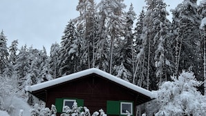 En træhytte med skråt tag, omgivet af sneklædte træer og et snefyldt hegn.