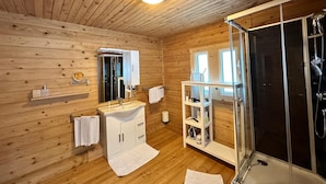 Et badeværelse i en træhytte med vask, spejl, håndklædestang, brusebad og et hyldesæt med toiletartikler.