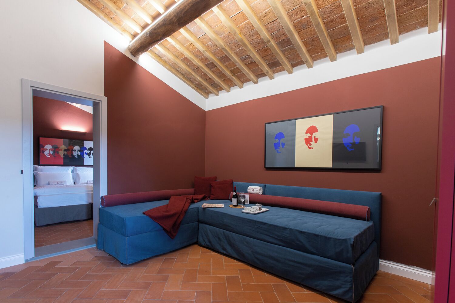 A room with a blue sofa, a bed with white bedding, a red wall, and a ceiling with exposed wooden beams.