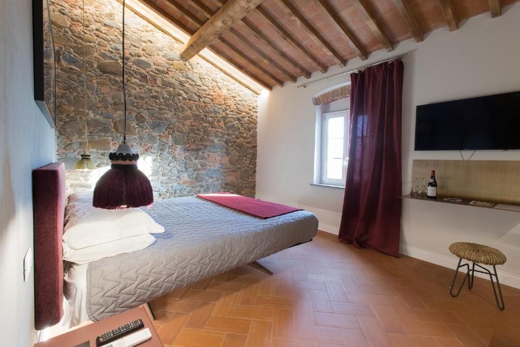 Un dormitorio con pared de piedra, vigas de madera en el techo, cama, televisor, ventana con cortinas y un taburete pequeño.