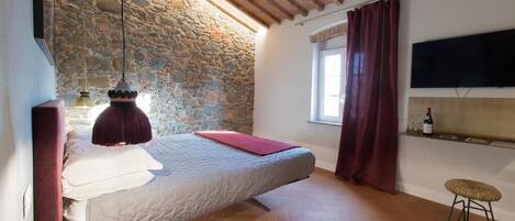Un dormitorio con pared de piedra, vigas de madera en el techo, cama, televisor, ventana con cortinas y un taburete pequeño.