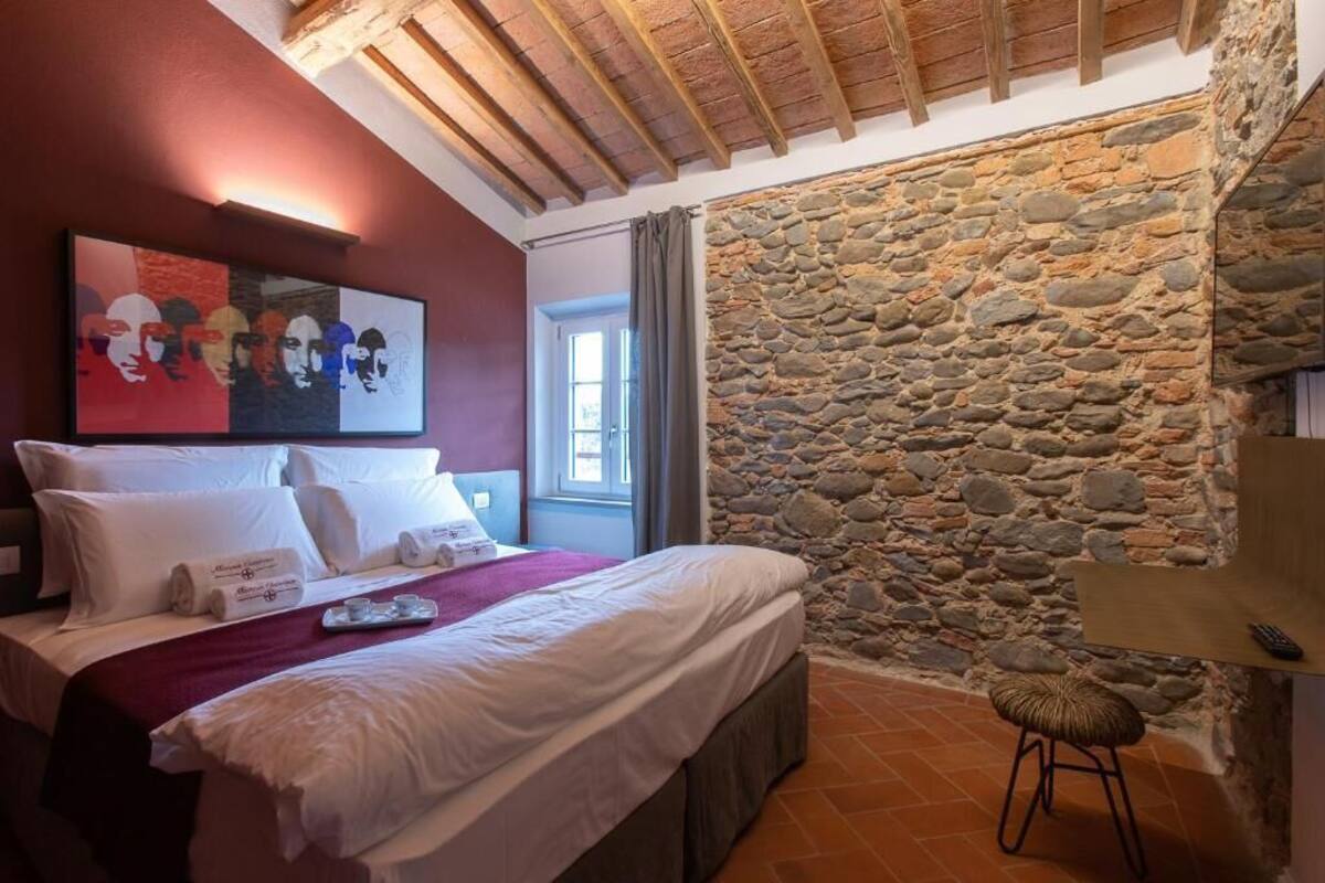 A bedroom with a stone wall, a bed with white linens, a painting on the wall, and a window with curtains.