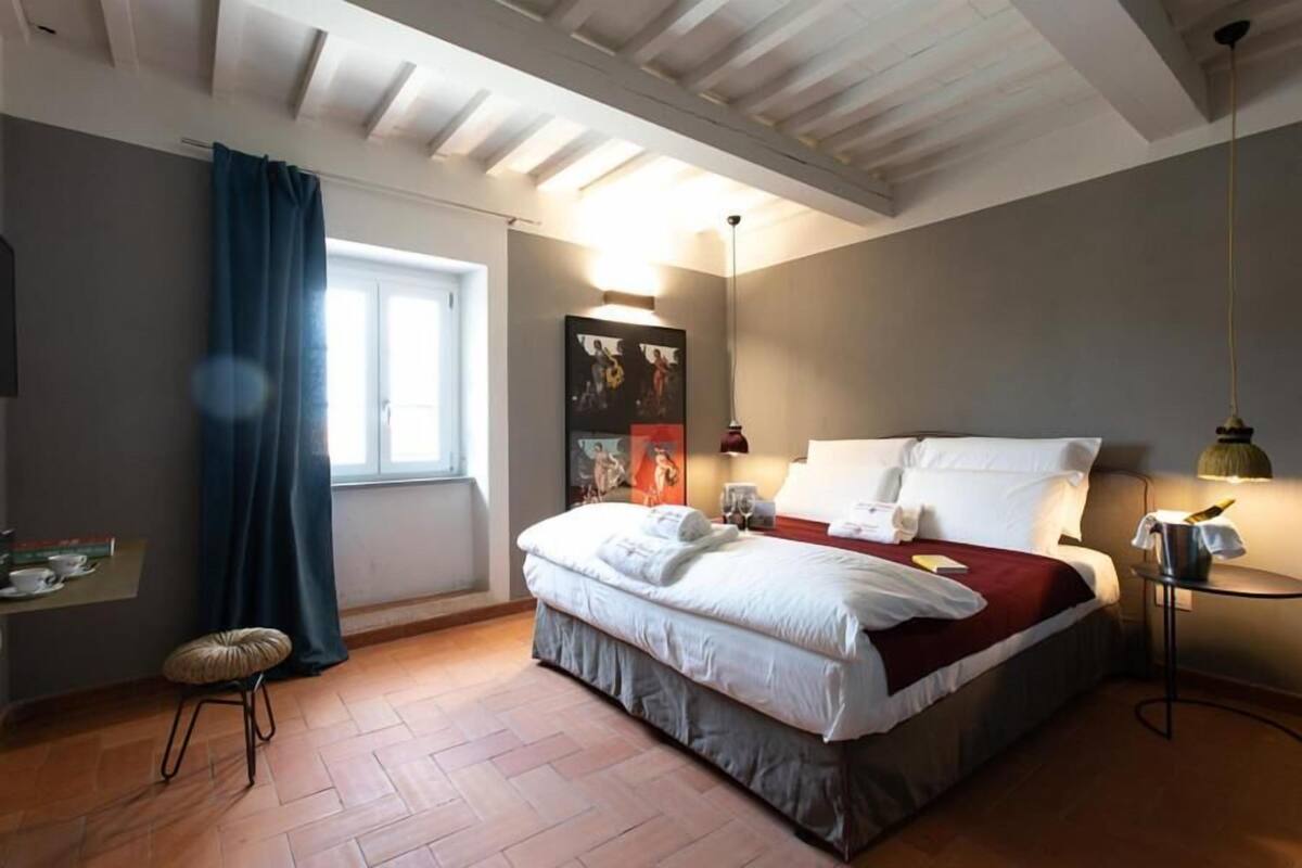 A bedroom with a bed, a bedside table, a lamp, a window with curtains, and a wall-mounted artwork.