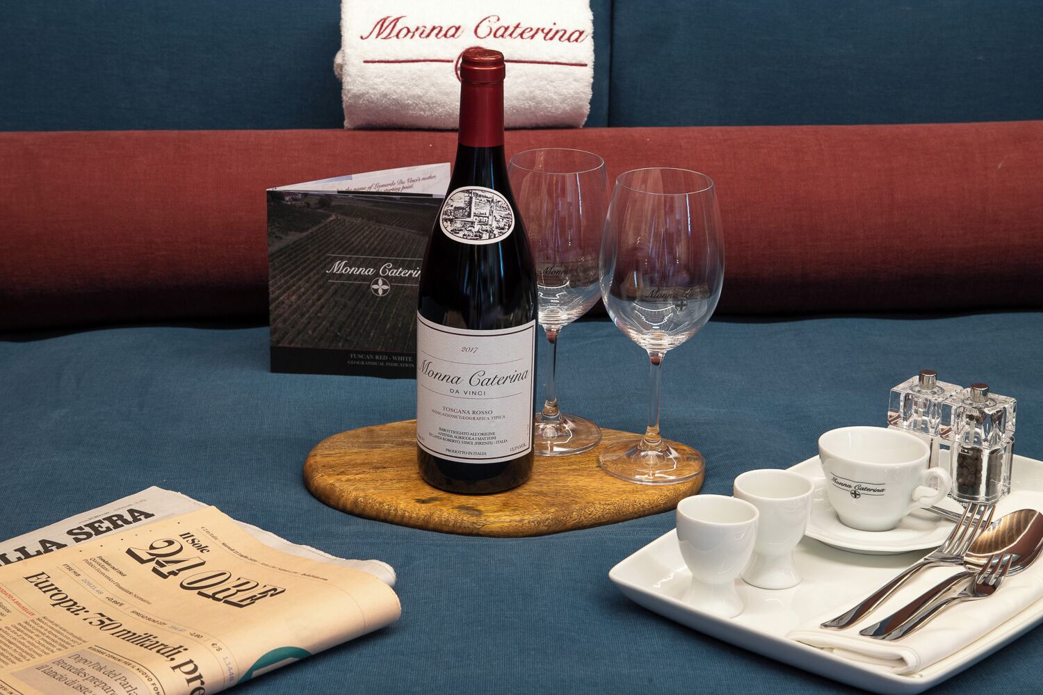 A bottle of wine, two glasses, a tray with cups and cutlery, and a newspaper on a blue bedspread.