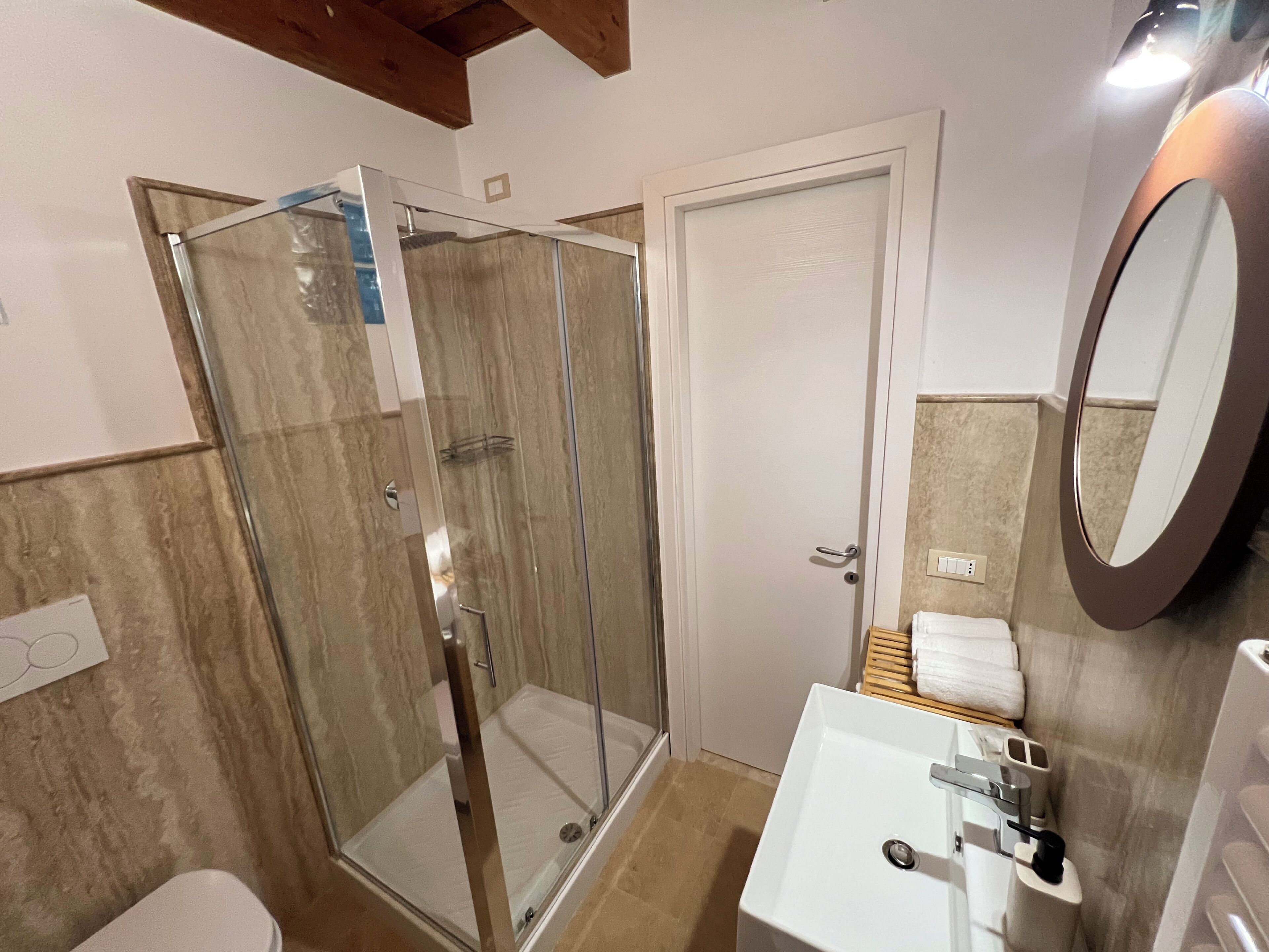 A bathroom with a glass shower enclosure, a white sink with a mirror, and a wooden shelf with towels.