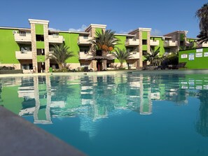 Una piscina con il riflesso di edifici, circondata da palme e un cielo sereno.