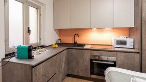 Eine kompakte Küche mit Mikrowelle, Spüle und Backofen, eine türkisfarbene Kaffeemaschine und ein Fenster mit Jalousien.