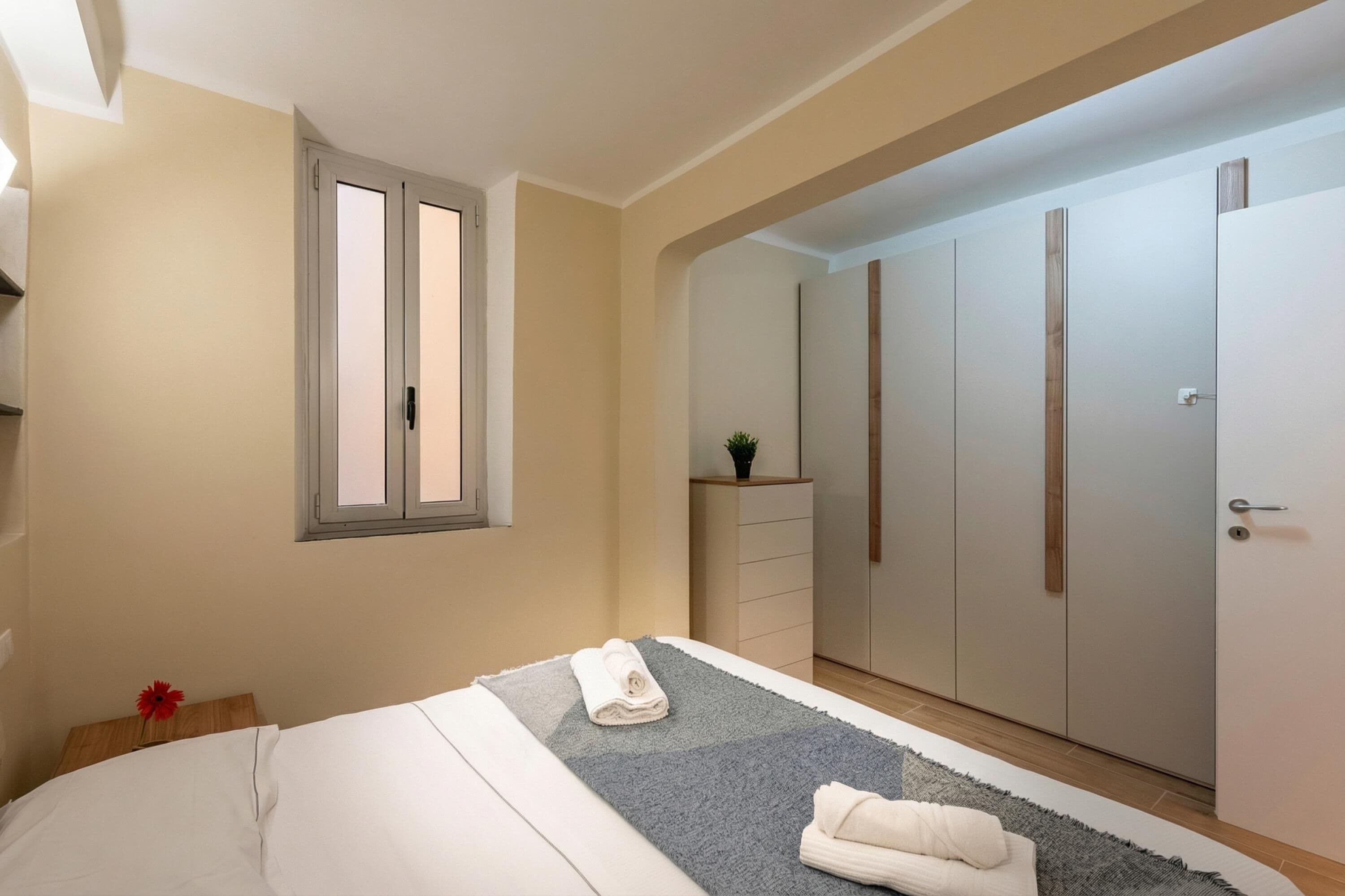 A bedroom with a bed, a bedside table with a flower, a window, a wardrobe, and a small plant on a dresser.
