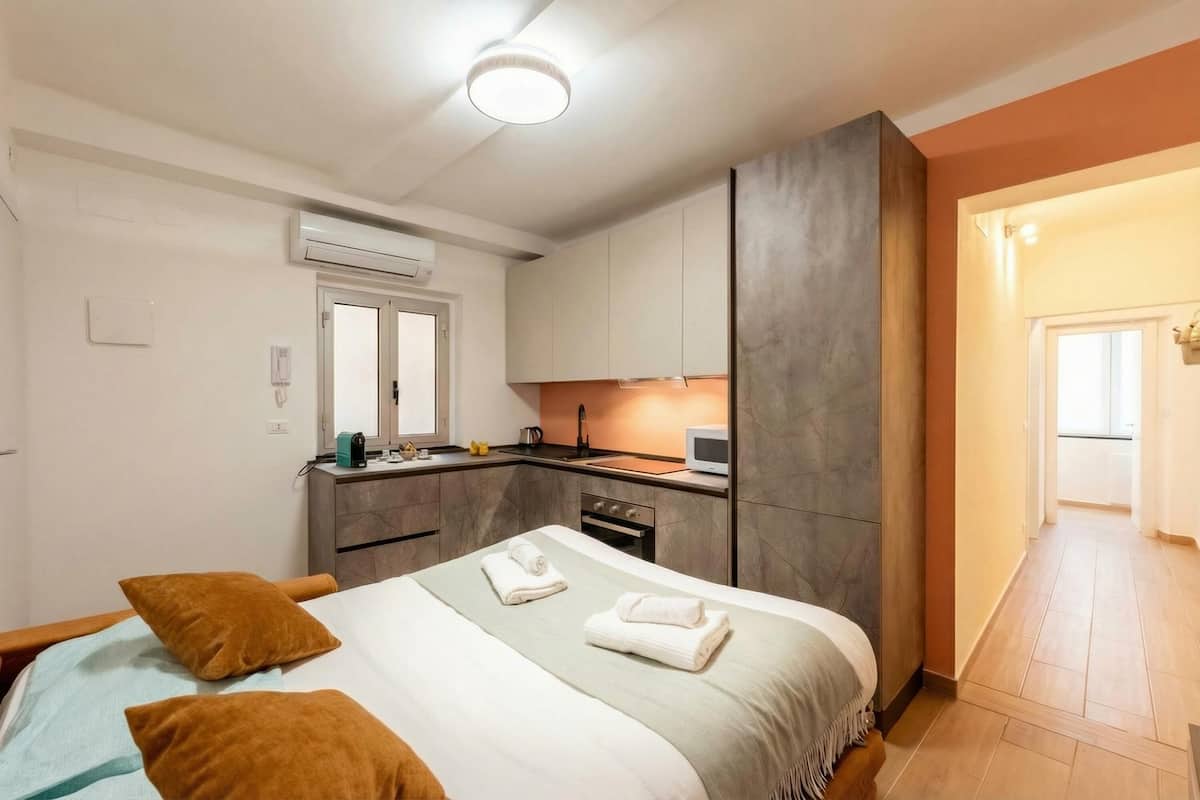 A bedroom with a bed, pillows, a bedside table, a built-in wardrobe, a kitchen area with a microwave, and a bathroom with a sink.