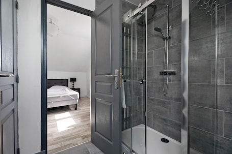 A modern bathroom with a glass-enclosed shower, a bathtub, and a visible bedroom with a bed and nightstand.. Gîtes des Lacs - Le Muscat: Shared Pool, Sauna & Jacuzzi, Private Terrace, Wi-Fi