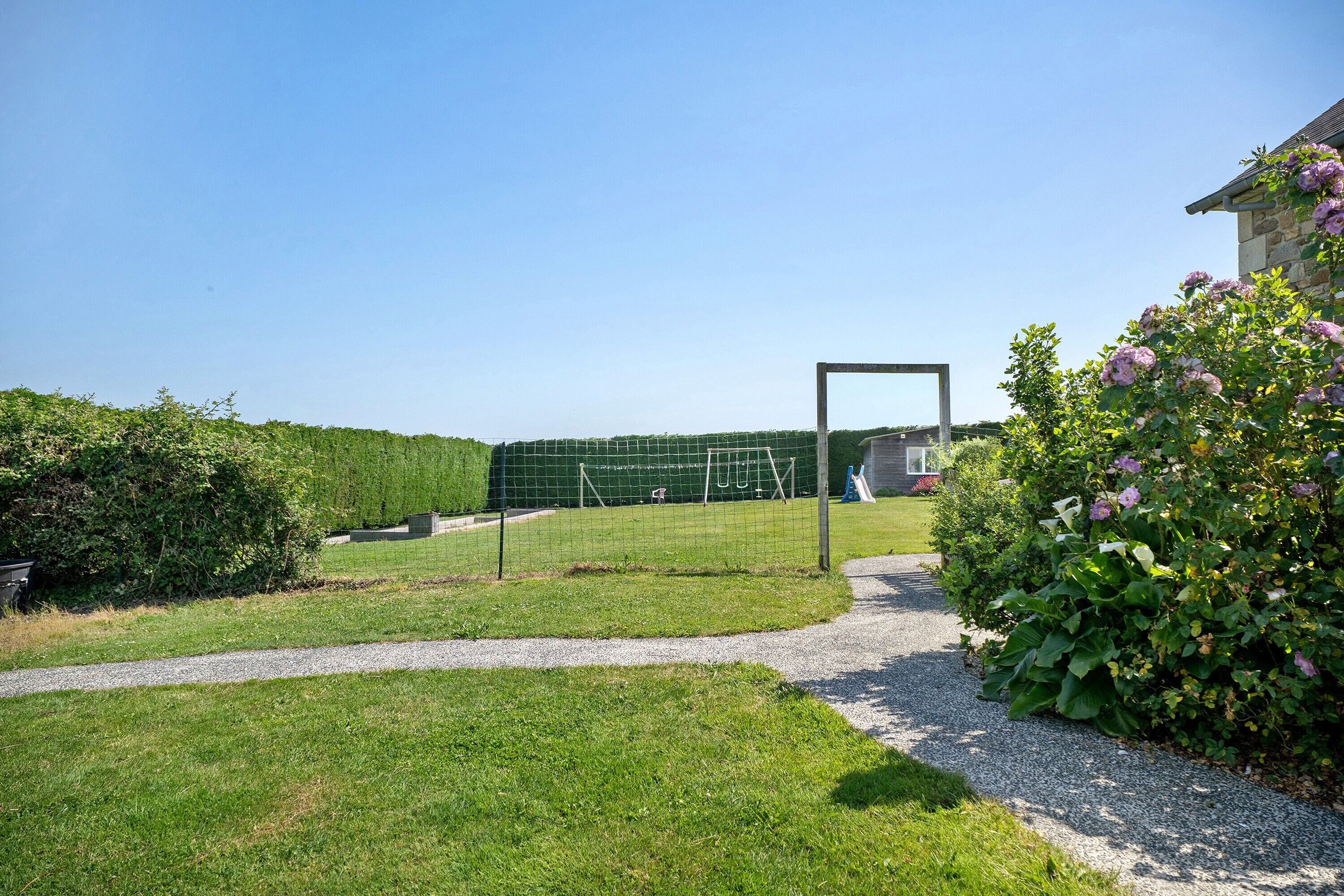 A grassy outdoor area with a pathway leading to a house, a fenced sports area, and a bush with flowers.