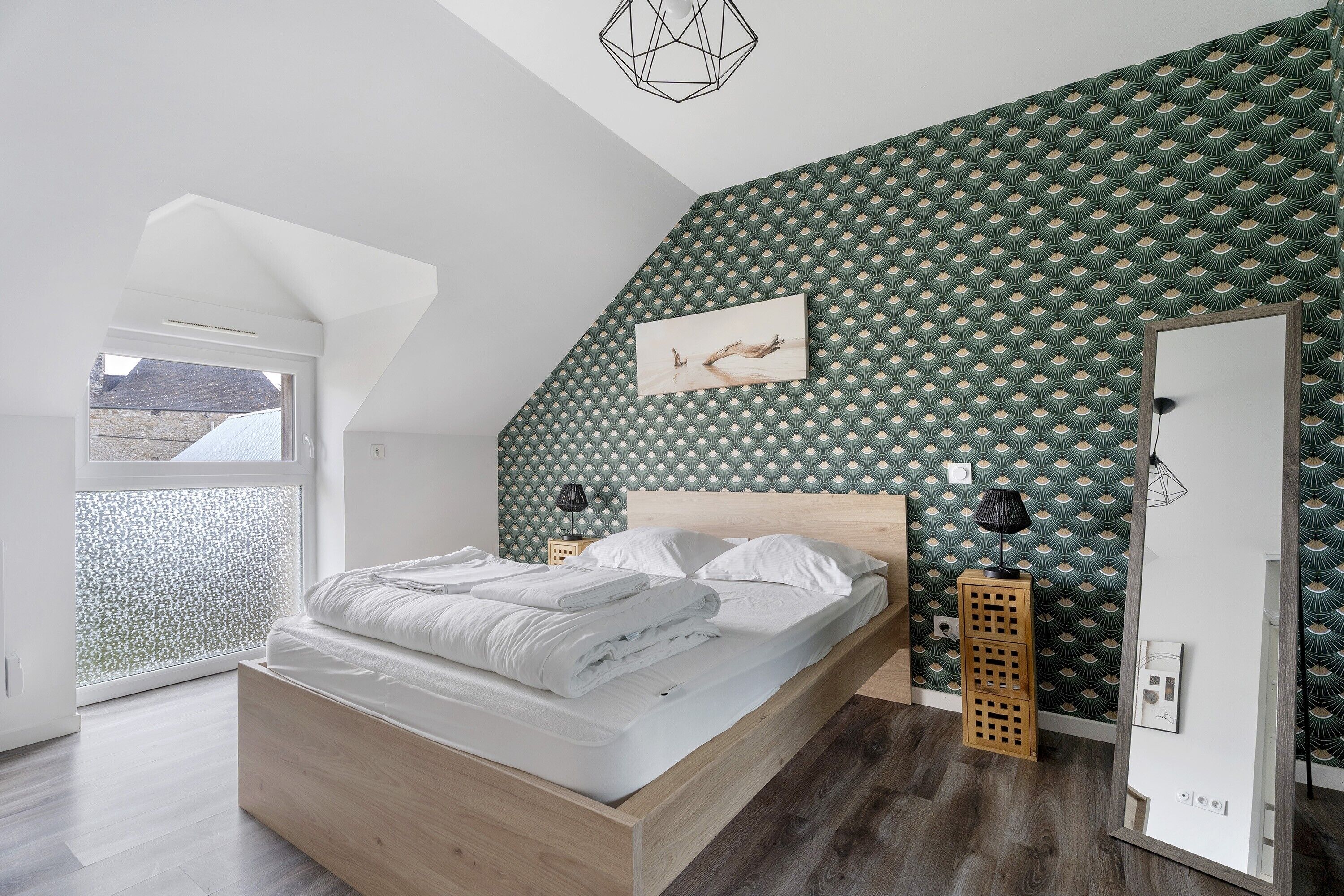 Une chambre avec un plafond en pente, un lit recouvert de linge de lit blanc, un papier peint à motifs, un tableau encadré accroché au mur et une fenêtre offrant une vue.