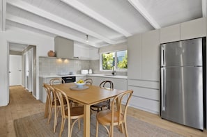 A modern kitchen with white cabinetry, a stainless steel refrigerator, a dining table with chairs, and a window with a view of greenery. - Piccolo Casa di Rye (Melbourne)
