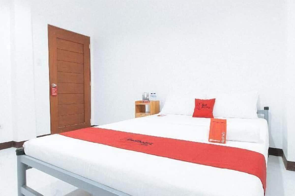 A single bed with a white and red bedspread, a wooden door, and a small bedside table with a lamp.