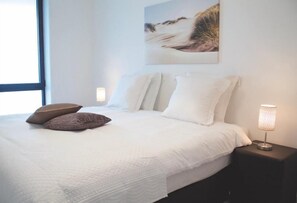 Una cama bien tendida con ropa blanca y dos almohadas marrones, una lámpara de noche y un cuadro en la pared.