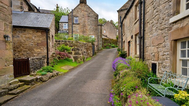 A narrow stone street with traditional stone buildings, a metal bench, and a small garden with flowers. - East Bank (Matlock)