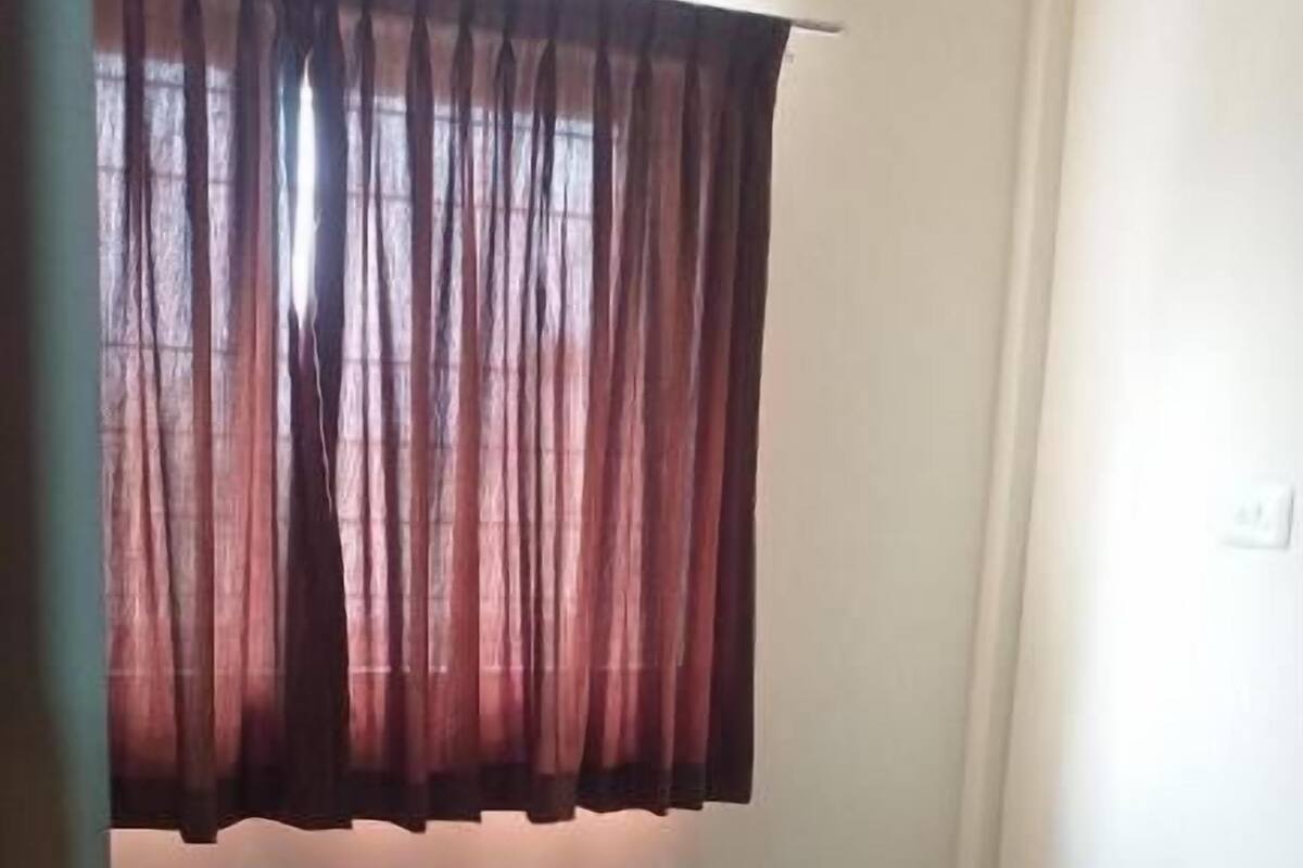 Ein Einzelbett mit weißer Matratze und braunem Kopfteil, ein Fenster mit dunklen Vorhängen sowie eine Klimaanlage darüber.