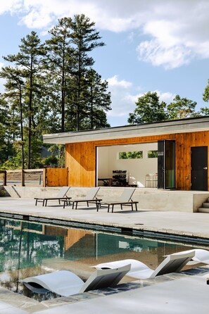 Un espace piscine extérieur moderne, doté de chaises longues, d’une terrasse en bois et d’un bâtiment aux grandes fenêtres.