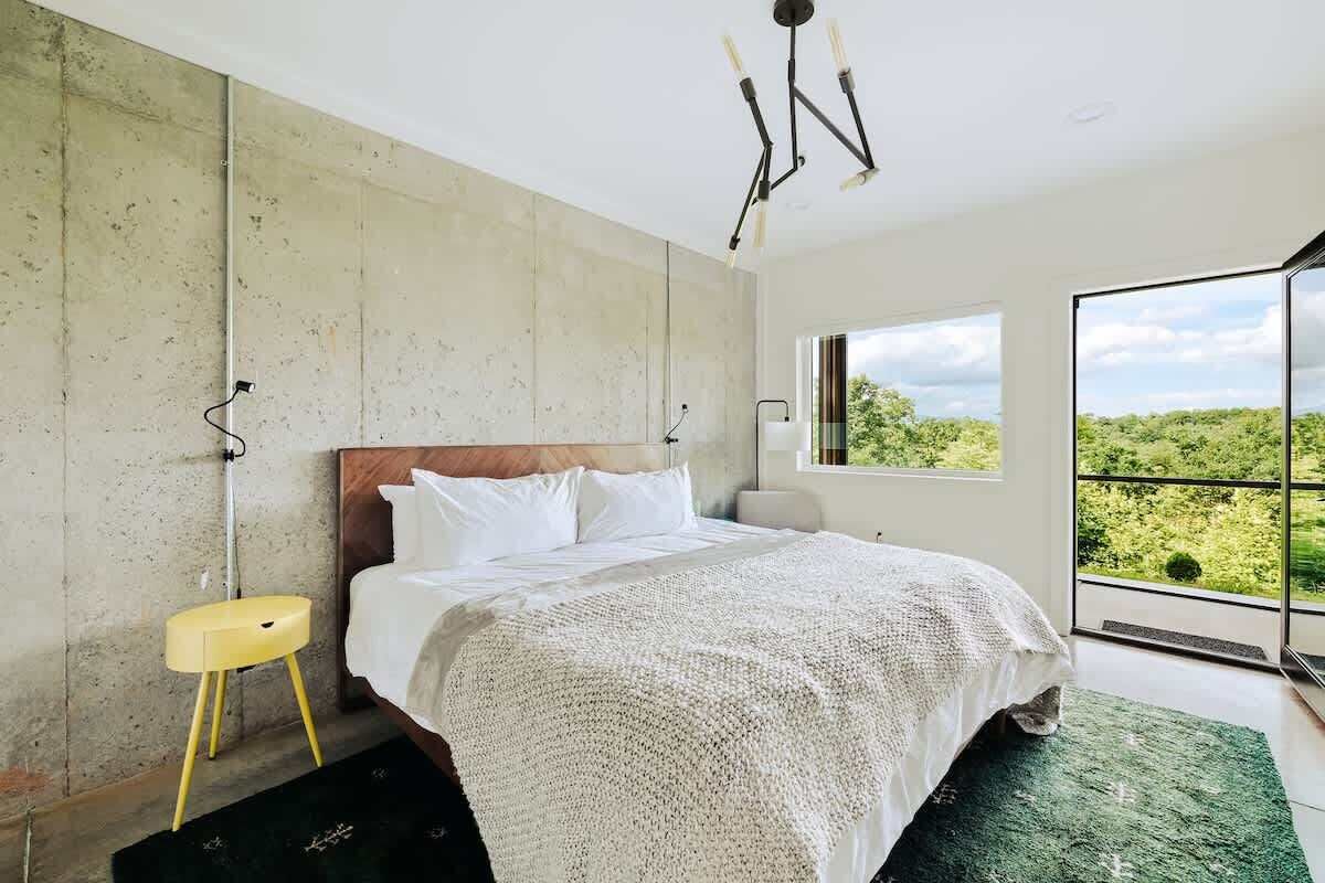 Une chambre avec un grand lit, une vue sur la verdure par la fenêtre et un mur en béton.