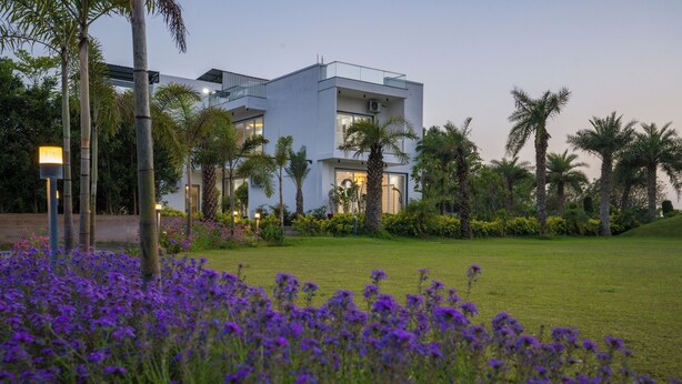 A modern building with a flat roof, surrounded by a well-maintained lawn and palm trees. - ELIVAAS Adhiva Retreat 5 BHK Farmhouse (Gurugram)