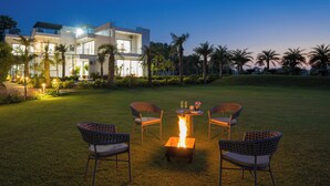 A modern building with a fire pit and chairs in the foreground, surrounded by a well-maintained lawn and palm trees. - ELIVAAS Adhiva Retreat 5 BHK Farmhouse (Gurugram)