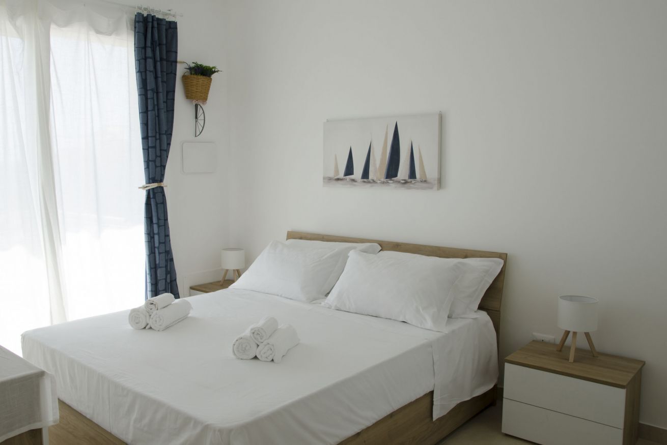 A neatly made bed with white linens and rolled towels, a bedside table with a lamp, a wall-mounted plant, and a painting of sailboats.