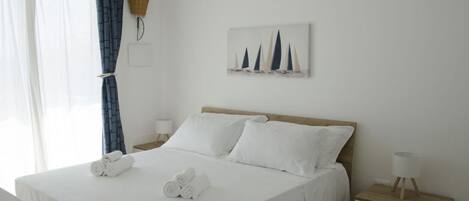 A neatly made bed with white linens and rolled towels, a bedside table with a lamp, a wall-mounted plant, and a painting of sailboats.