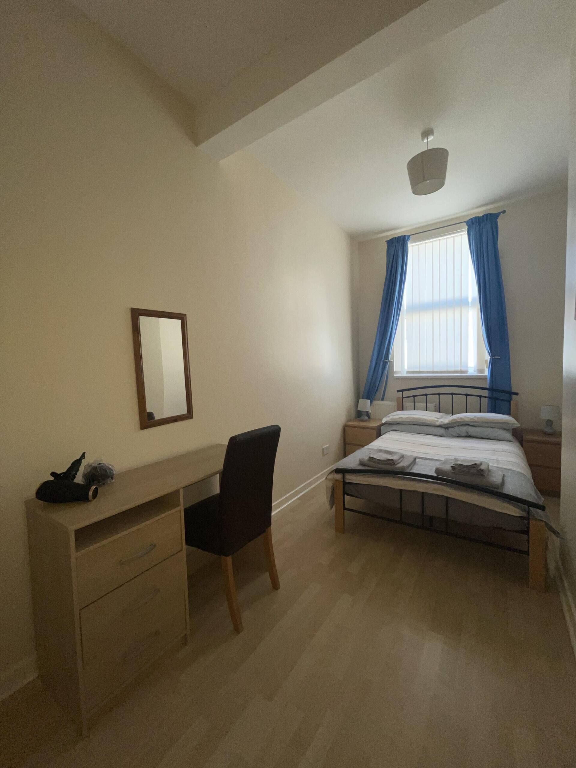 Ein Schlafzimmer mit Bett, Schreibtisch, Stuhl, einem Fenster mit Vorhängen und einer Deckenleuchte.