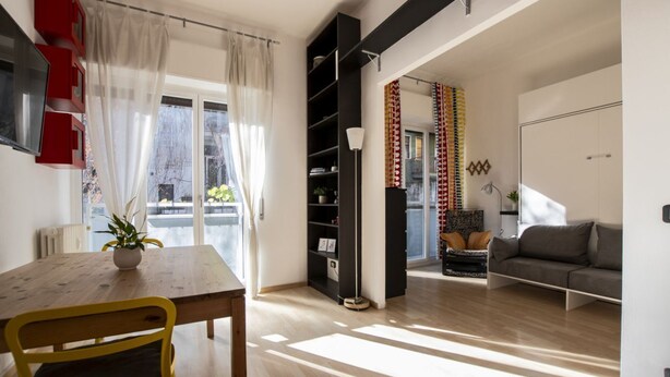 A modern living room with a dining area, a sofa, a bookshelf, and a large window with curtains. - Open Space Apartment near Lambrate Station (M2 and FS) & Politecnico (Milano)