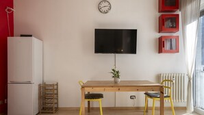 A room with a wooden table, two chairs, a TV, a clock, a radiator, and a window. - Open Space Apartment near Lambrate Station (M2 and FS) & Politecnico (Milano)