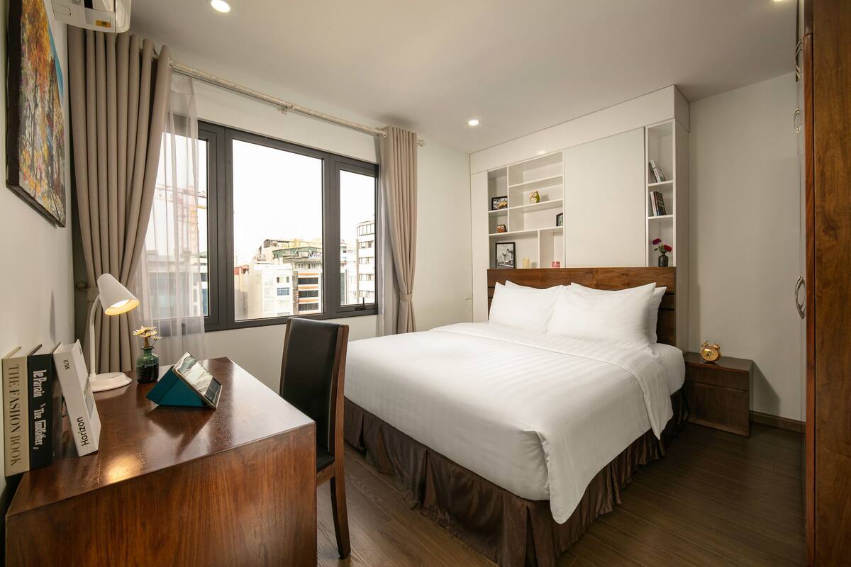 A hotel room with a bed, a desk with a lamp, a chair, a bookshelf, and a window with curtains.