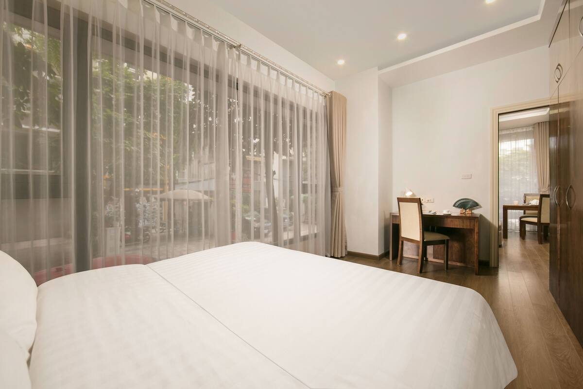A hotel room with a large bed, a desk, and a chair. There is a window with sheer curtains and a view of greenery outside.