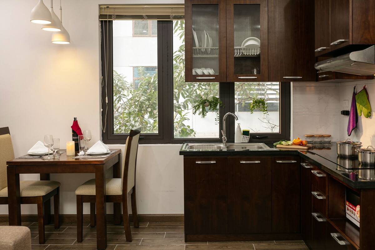 A modern kitchen with dark wood cabinets, a window with a view of greenery, a dining area with a table set for two, and a countertop with kitchen appliances.