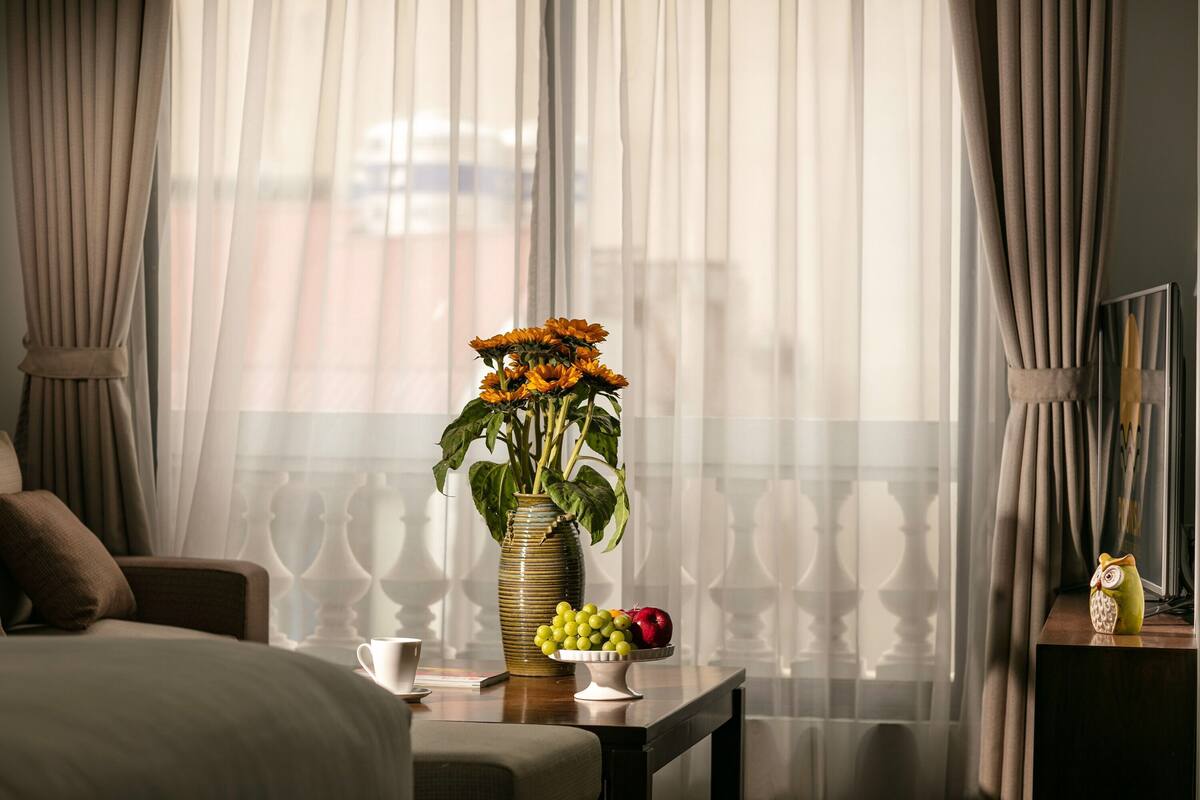 A hotel room with a bed, a bedside table with a vase of flowers, a bowl of fruit, and a cup.