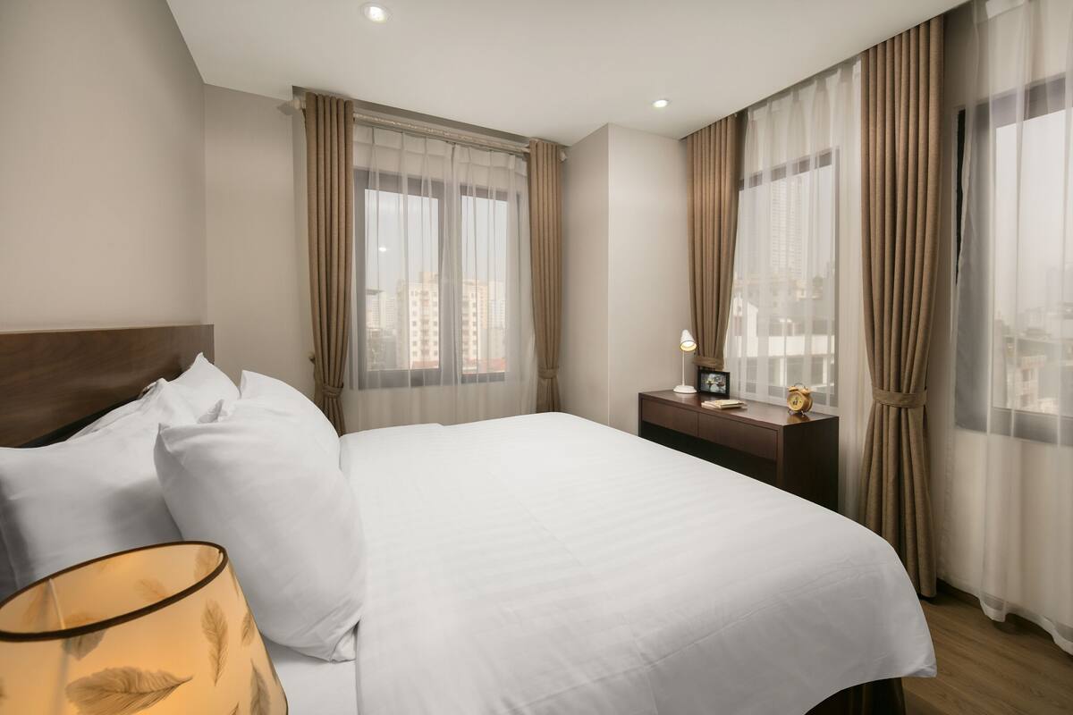 A hotel room with a large bed, a bedside table with a lamp, a nightstand, and a view of the city through sheer curtains.