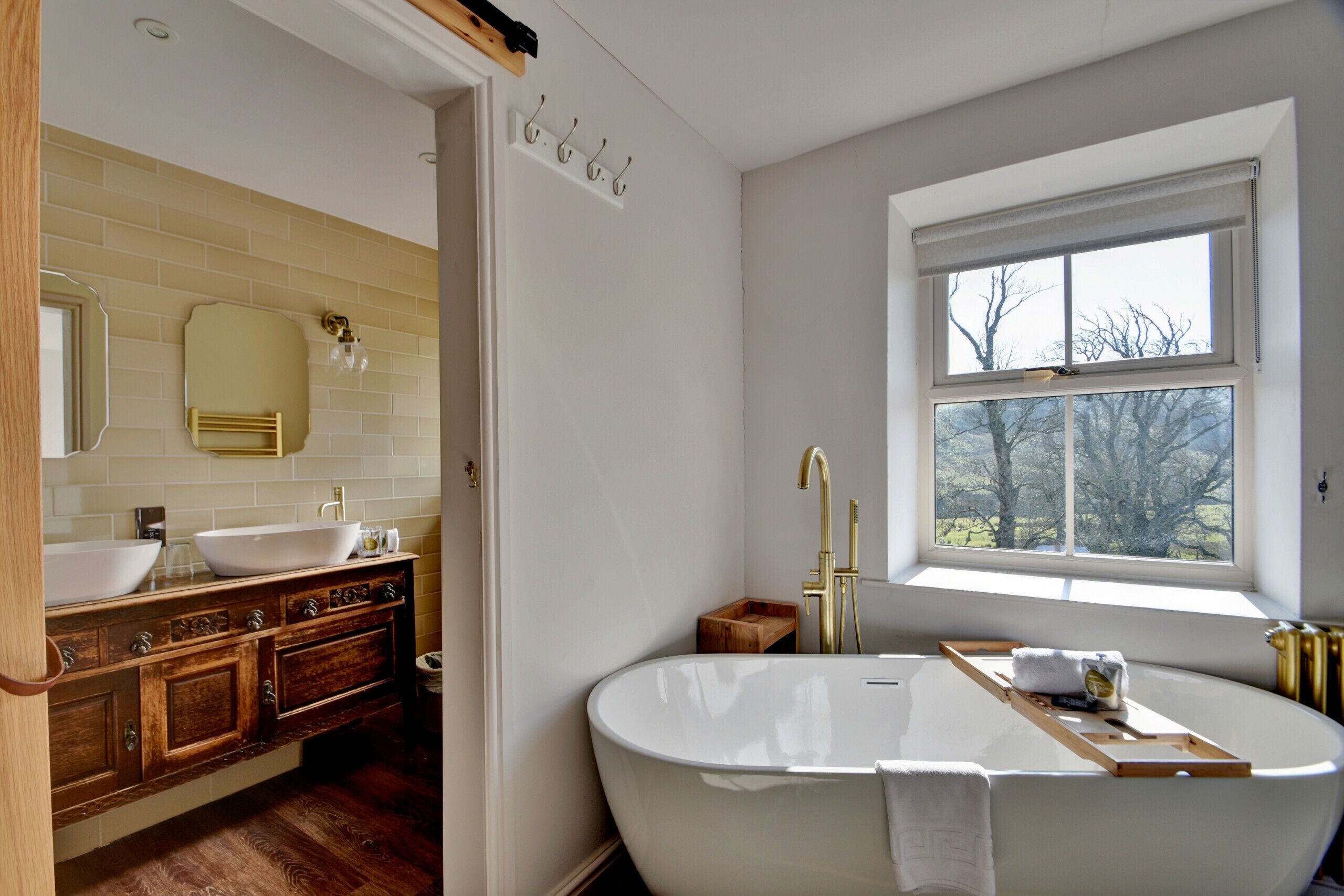 A modern bathroom with a freestanding bathtub, two sinks, wooden cabinetry, and a window with a view.