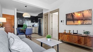 A modern living room with a sofa, a table with a plant, a wooden cabinet, and a kitchen in the background. - NOK Modern 1BR With Terrace by Vistacana Rentals (Punta Cana)