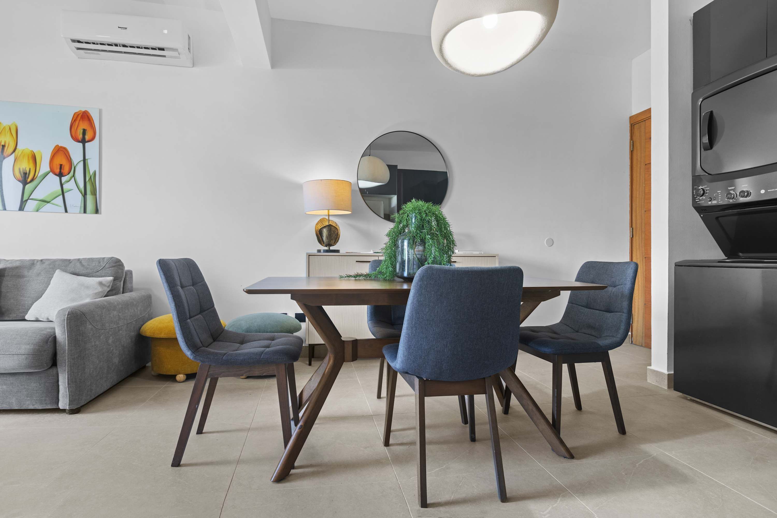 A modern living room with a dining table, chairs, a sofa, a lamp, a mirror, and a kitchen area with a stove.