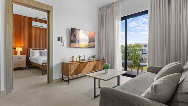 A modern hotel room with a bed, a sofa, a small table, a cabinet, and a large window with curtains. - NOK Modern 1BR With Terrace by Vistacana Rentals (Punta Cana)