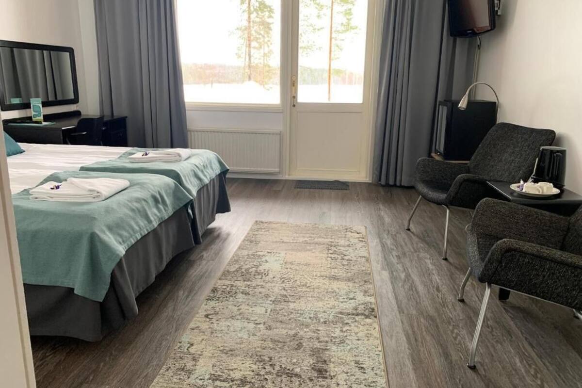 A hotel room with a bed, a desk, a chair, a television, and a view of trees through the window.