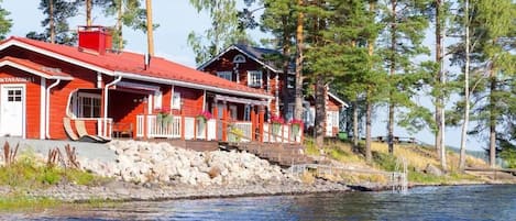 Une cabane en bois rouge, dotée d’un porche, située au bord d’un lac, avec des arbres en arrière-plan.